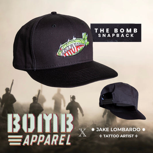 The image shows a black 9FIFTY snapback cap with The Original Atom Bomb logo embroidered in the center, designed by Jake Lombardo. The cap is displayed in two different angles, one showing the front view with the logo, and the other showing the back view with a subtle 'BOYB' logo on the back panel.