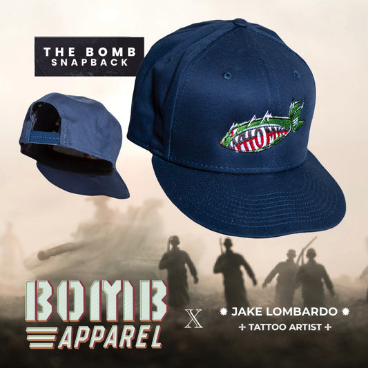 The image features a navy-colored 9FIFTY snapback cap with The Bomb logo embroidered at the front. The cap is displayed in two views, one showing the front and the other showing the back, with a caption that reads 'THE BOMB SNAPBACK' and 'JAKE LOMBARDO + TATTOO ARTIST'.
